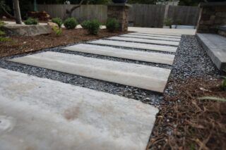 Wide concrete pavers form a path with dark gravel between them, bordered by mulch, small plants, and a wooden fence in the background.
