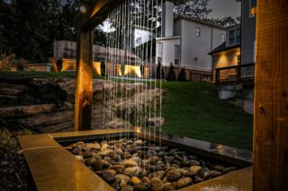 A modern backyard water feature with streams cascading onto smooth river rocks, set near a house at dusk in Atlanta.