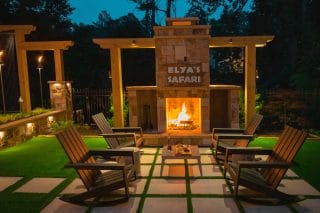 Nighttime patio with a fireplace labeled Elyas Safari, four wooden chairs, tiled floor, artificial grass, and wooden pergolas.