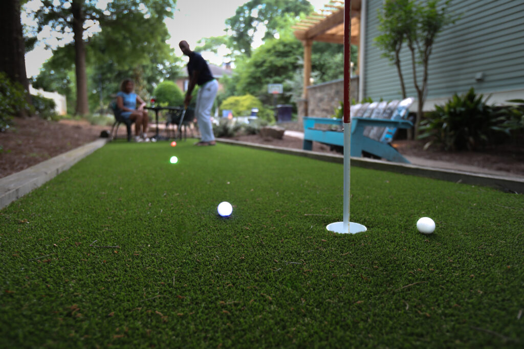 Two people play mini-golf on artificial turf in a backyard, with colorful golf balls near a flagged hole and trees and a house behind them.
