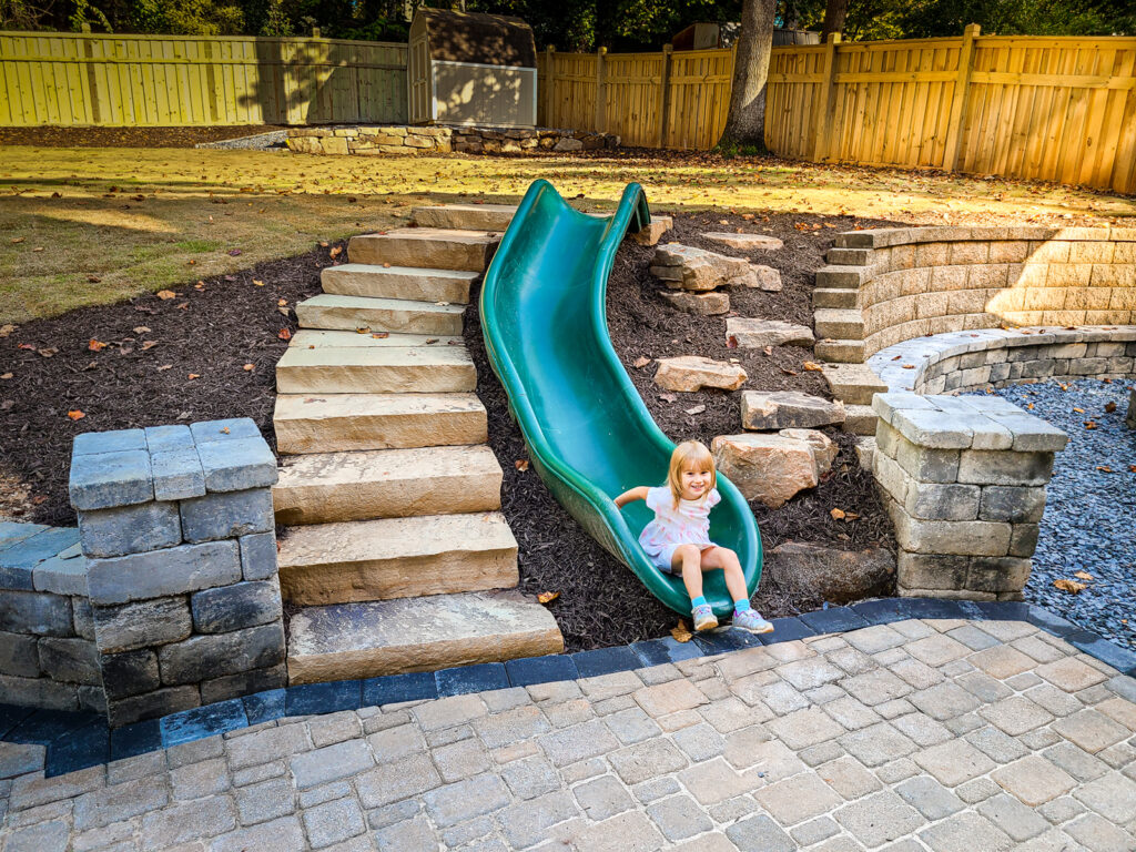 A young child in a pink dress sits at the bottom of a curved green slide in a landscaped backyard with stone steps and retaining walls.