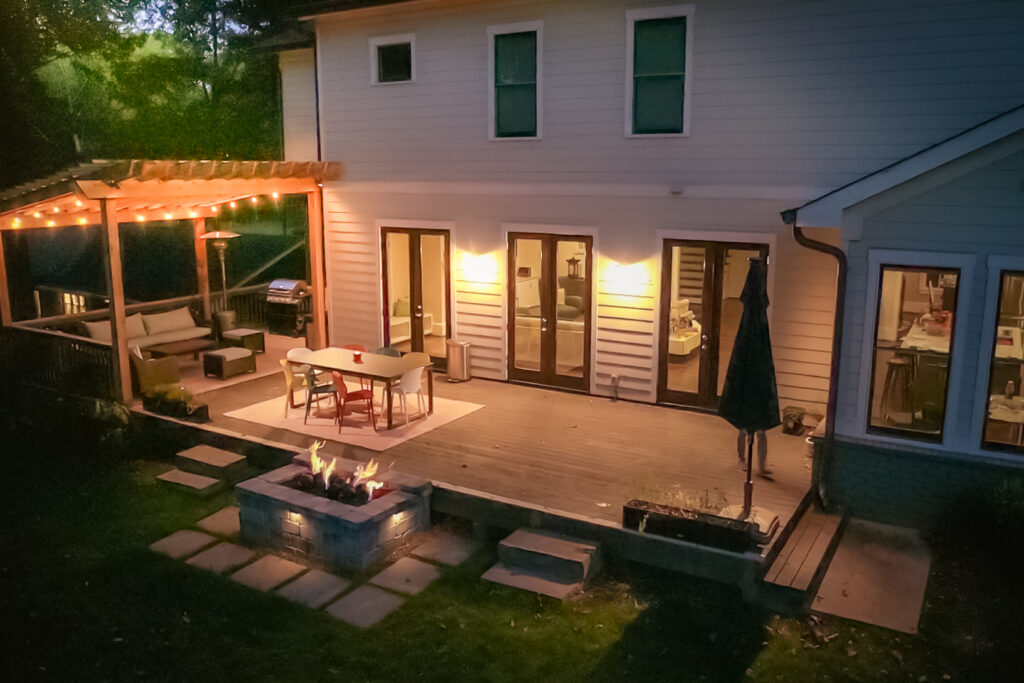 A backyard deck at dusk features string lights, a dining table, fire pit, outdoor seating, and a grill under a pergola by a white house.