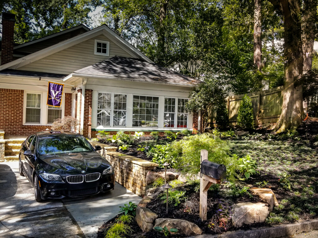 A black car sits in the driveway of a brick house with large windows, surrounded by a neat yard, stone borders, and a wooden mailbox.