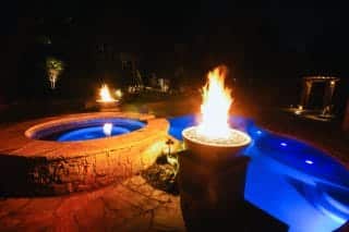 A backyard pool and hot tub glow at night with blue underwater lights and two fire bowls, creating a dramatic, inviting scene.