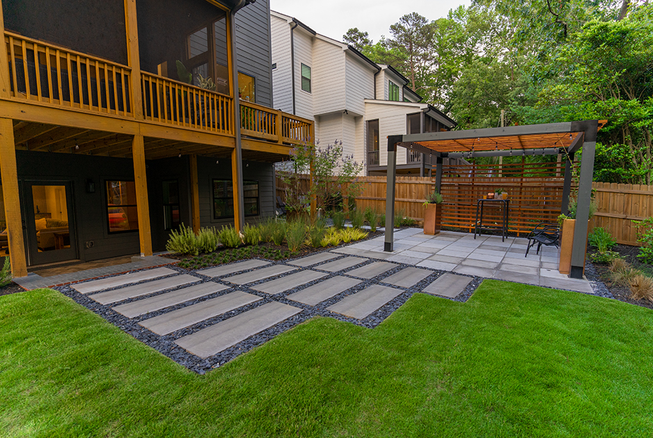 A modern backyard features a patio with a pergola, paver stones and gravel, green lawn, plants, and a wooden fence by two multi-story houses.