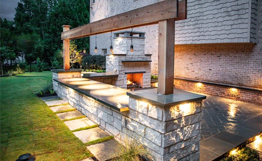 A modern patio at dusk features a stone fireplace, hanging lights, wooden beam, and stone pillars, surrounded by greenery and a stone walkway.