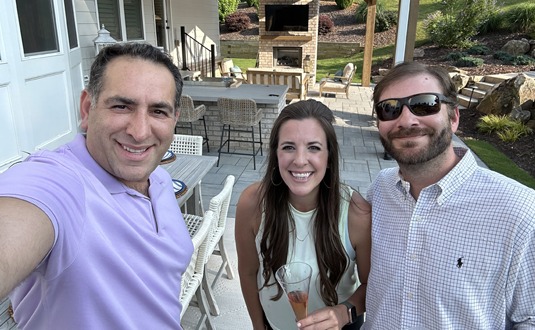 Three people smiling for a selfie on a sunny patio with outdoor furniture and a fireplace; one woman holds a drink, one man wears sunglasses.
