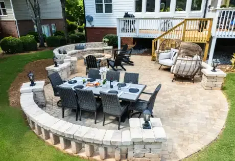 A backyard patio with stone hardscaping has a dining set, curved seating wall, Adirondack chairs by a fire pit, and cushioned egg chair amid grass and shrubs.