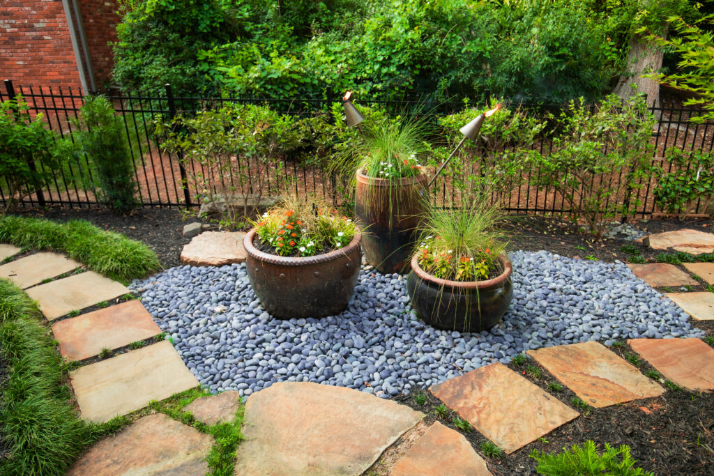 Three large flower pots with tall grasses and flowers rest on gray pebbles among stone pavers and greenery in a backyard with a black metal fence.