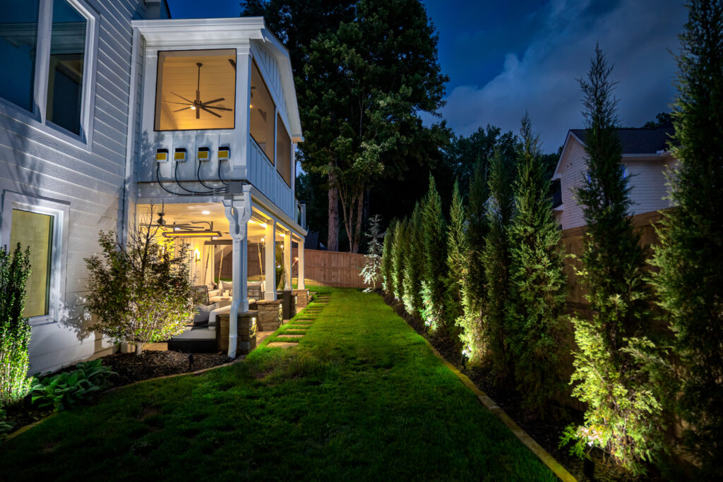 A modern house at night with a covered patio, outdoor seating, ceiling fan, and backyard lights illuminating tall trees on a green lawn.