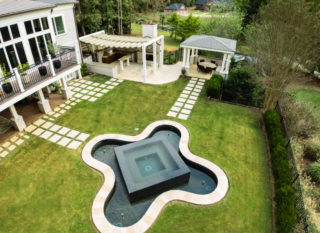 Aerial view of a backyard featuring a geometric pool, green lawn, stone paths, patio with pergola, outdoor kitchen, gazebo, and surrounding trees.