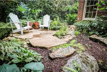 Two white Adirondack chairs on a stone patio are surrounded by lush greenery, potted plants, rocks, and a brick wall with a window.