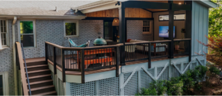 A covered patio deck attached to a house has a hammock, grill, dining table, TV, seating, black railings, and is surrounded by trees.