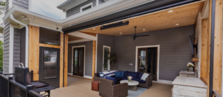 A modern covered patio has a wood-paneled ceiling, sectional sofa, coffee table, ceiling fan, grill, and decorative lights.