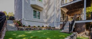 A modern gray house features a second-floor balcony, artificial grass lawn, stone wall, landscaped plants, and concrete stairs up the slope.