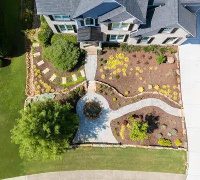 Aerial view of a landscaped front yard with stone paths, green grass, shrubs, mulch beds, and a small circle in front of a gray house with white trim.