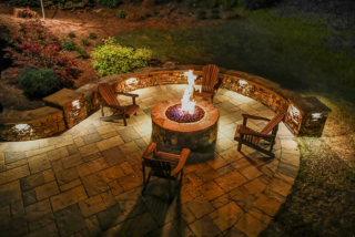 An elevated evening view shows chairs around a circular fire pit within curved stone seating walls and illuminated planting beds.
