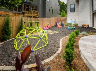 A fenced backyard has a green climbing dome, toy cars, a small slide, playhouse, and wagon on mulch beside curved garden beds and pathways.