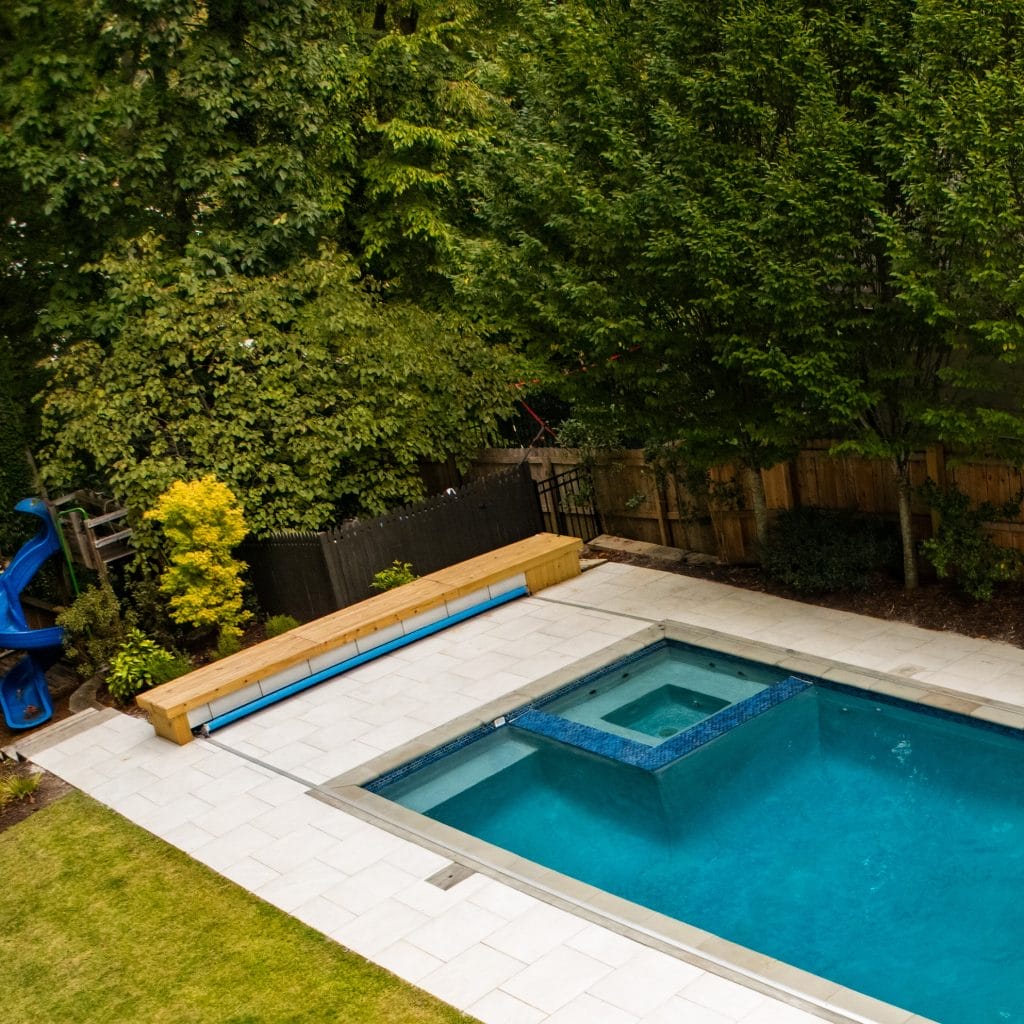 An elevated view shows a pool with a square spa beside a pale deck, wood fence and tall evergreen trees.