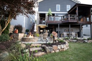 A landscaped backyard has a stone fire pit with Adirondack chairs, a metal sculpture, garden beds, and a gray house with an upper deck.