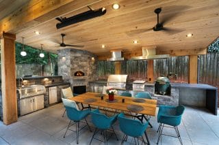 A covered outdoor kitchen with grills, pizza oven, sink, fridge, and a modern dining table with blue chairs under fans and wood ceiling.