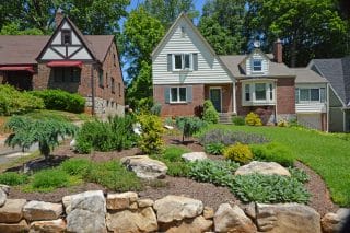Two brick houses with steep roofs sit amid landscaped yards of rocks, shrubs, and grass under a sunny blue sky with trees behind them.