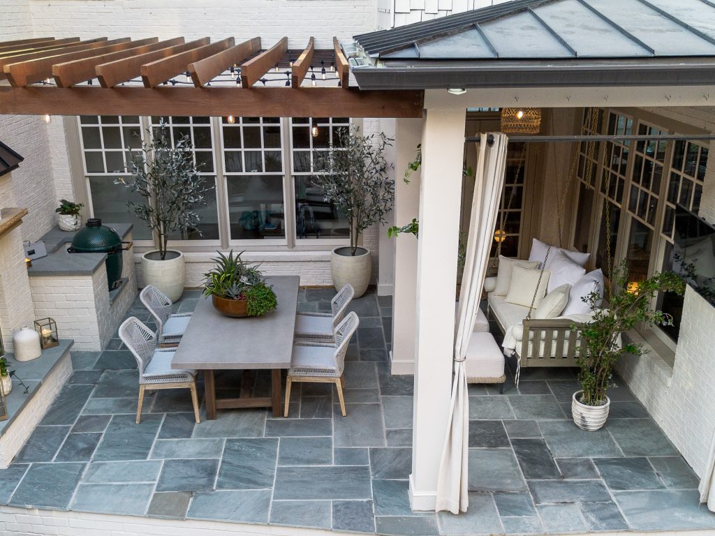 A modern patio features a pergola, dining set for six, potted plants, swing bench with cushions, gray stone floor, white brick walls, and large windows.