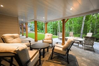 A covered patio with cushioned chairs, a round table, stone tile flooring, and views of a green backyard with trees and a fire pit.