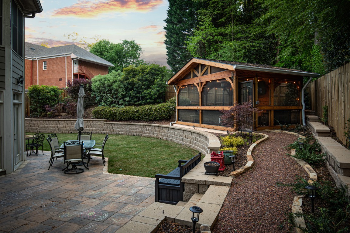 A backyard at sunset features a paved patio with seating, umbrella, fire pit, and a curved path to a wooden screened gazebo amid grass and trees.