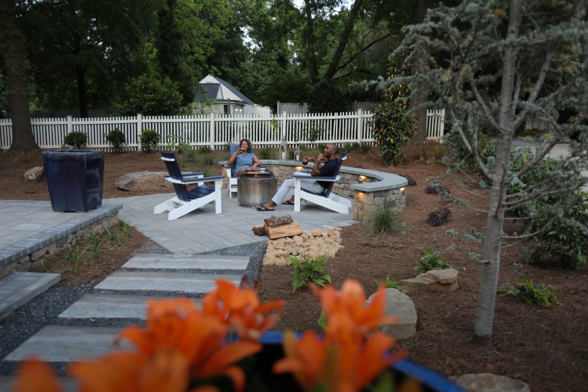 Two people sit by a fire pit on a stone patio surrounded by trees, flowers, orange blooms in the foreground, and a white fence with a house behind.