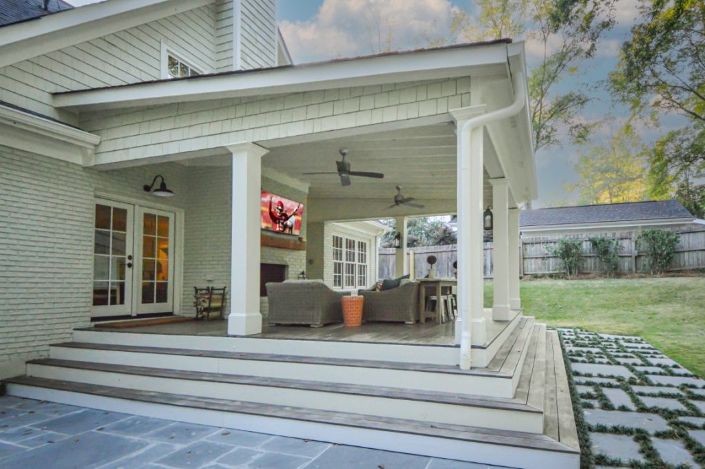 Covered backyard patio with ceiling fans, outdoor furniture, a wall-mounted TV, and steps to a grassy yard beside a light gray brick house.