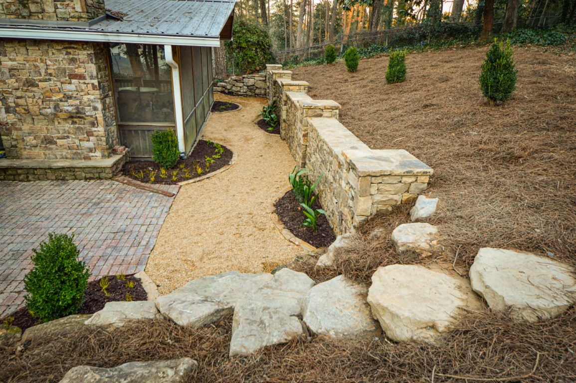 A stone house with a gravel path, retaining wall, landscaped yard of mulch and shrubs, and pine straw areas bordered by trees and a fence.