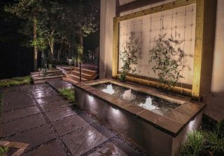 Modern outdoor garden at night features a lit rectangular water fountain, plants, stone path, wooden trellis wall, and steps leading to greenery.