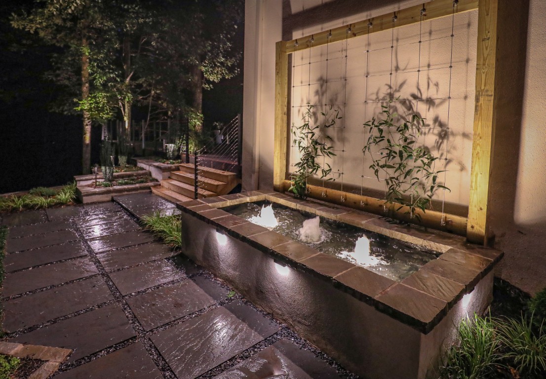 Modern outdoor garden at night features a lit rectangular water fountain, plants, stone path, wooden trellis wall, and steps leading to greenery.