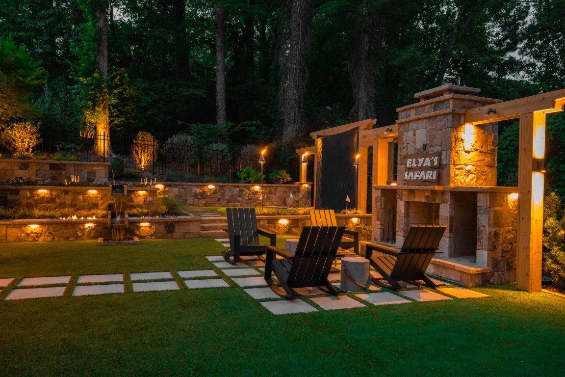 At dusk, four wooden chairs circle a fireplace on a cozy patio with string lights, stone pavers, green grass, and softly lit garden walls.