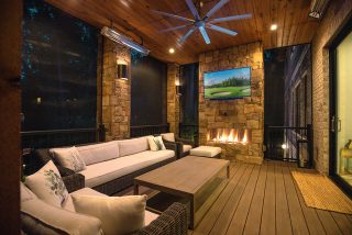 A covered patio with wicker seating, a wooden table, stone fireplace with TV, wood ceiling, fan, recessed lighting, and screened walls at night.