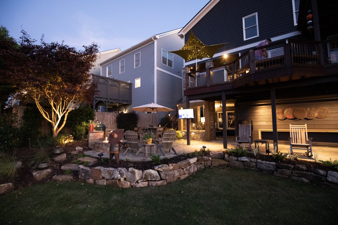 A cozy backyard at dusk features a firepit, Adirondack chairs, patio lights, stone landscaping, and a dark gray house with glowing windows.