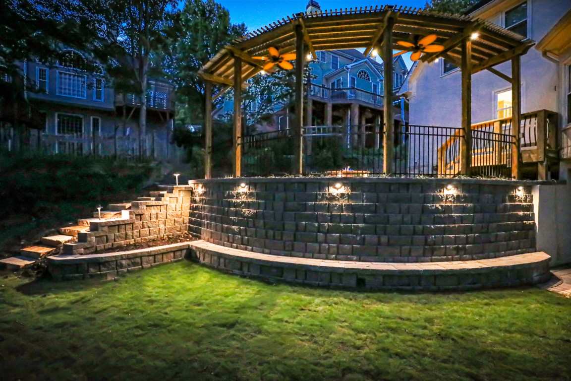 A backyard patio features a pergola, ceiling fans, accent lighting on stone walls, stone steps to a terrace, and grass with houses in the dusk background.