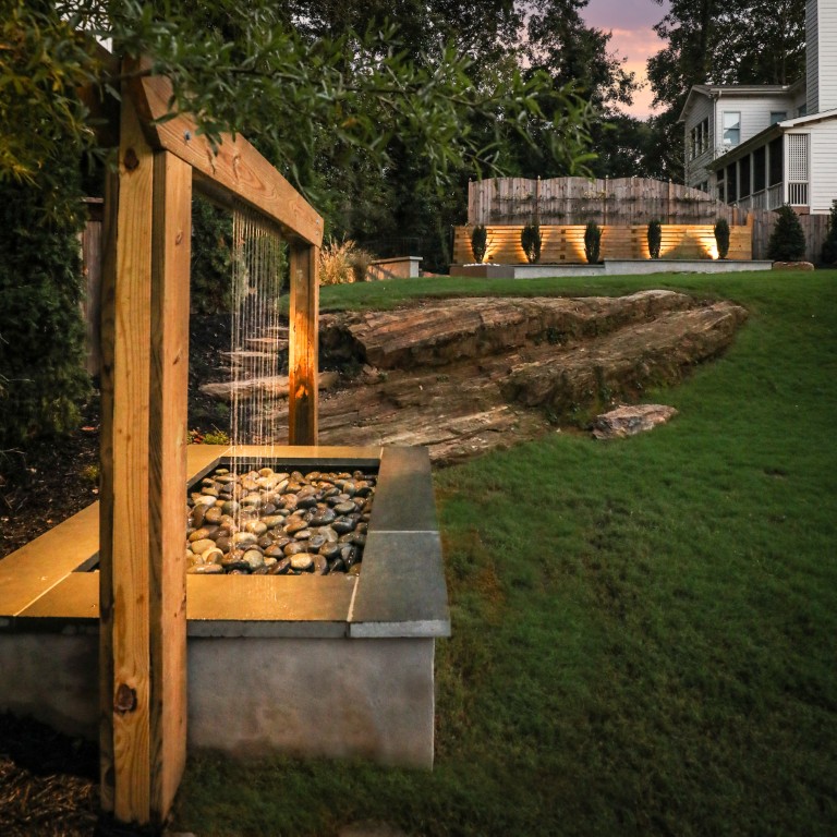 A twilight backyard has a modern wooden water feature with a waterfall, stone basin, grassy lawn, rock formation, and house with landscape lighting.