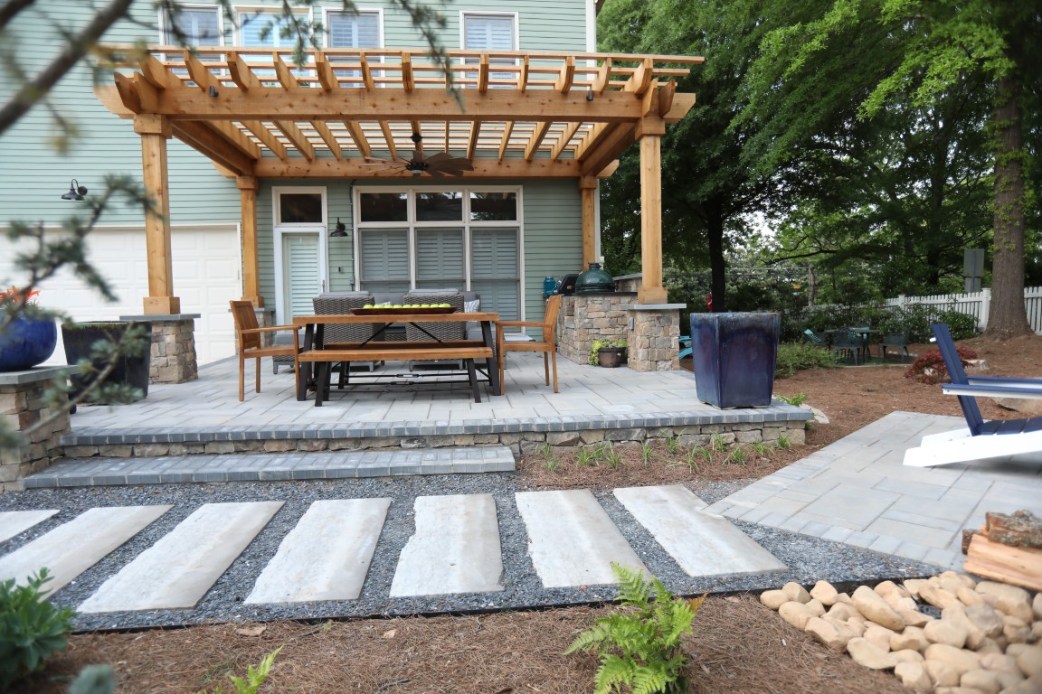 A backyard patio features a wooden pergola, dining set, paved walkway, blue chairs, and surrounding lush greenery.