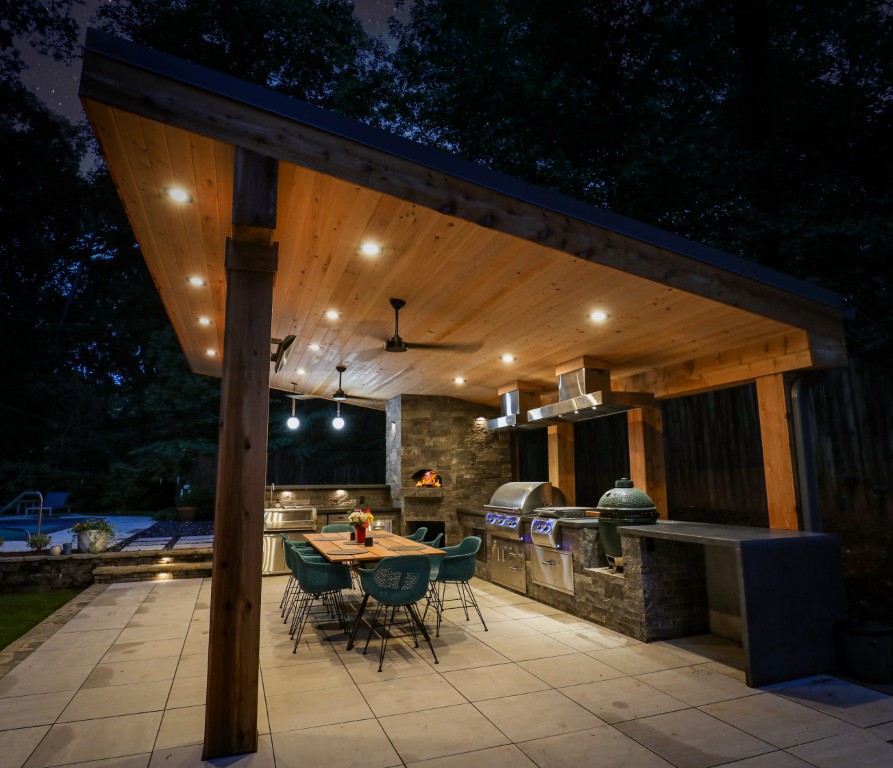 At night, an outdoor covered kitchen with a wooden ceiling, recessed lights, modern appliances, and teal dining chairs is warmly lit among trees.