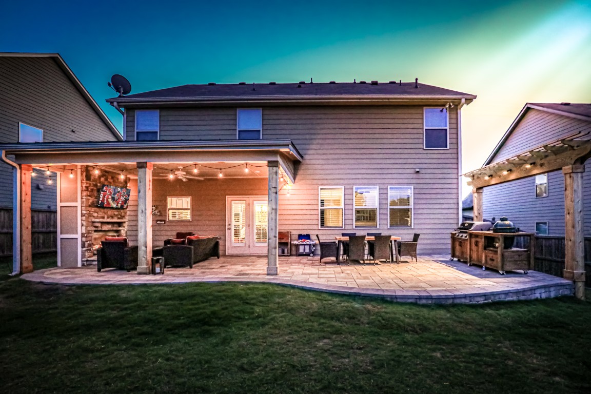 At sunset, a backyard patio with outdoor seating, string lights, a dining table, and a covered grill area sits beside a two-story house.
