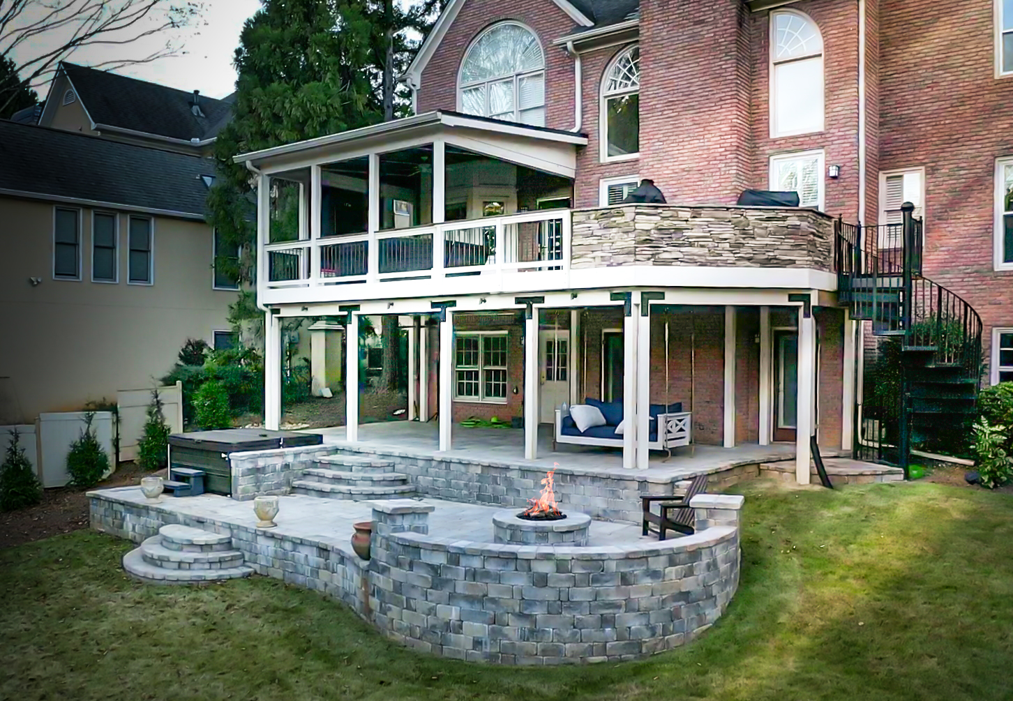 A two-story brick house features a large covered deck, patio with a stone fire pit, outdoor seating, a spiral staircase, and landscaped yard.