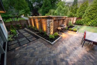 A modern backyard with stone and wood fencing, small garden, paver patio, grill, dining table, green lawn, and surrounding trees and shrubs.