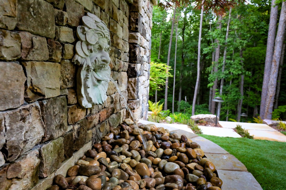 A stone wall fountain with a sculpted face spouts water onto smooth rocks, surrounded by green trees and a landscaped garden.