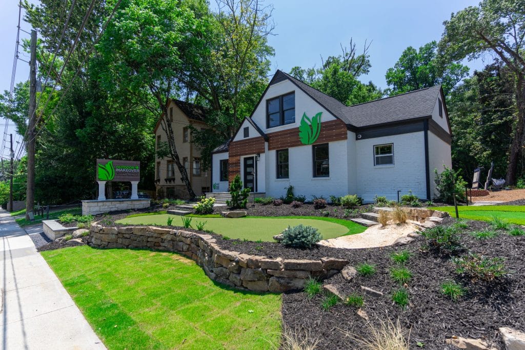 A white one-story building with a leaf logo has a landscaped front yard and a matching wooden sign by the street.