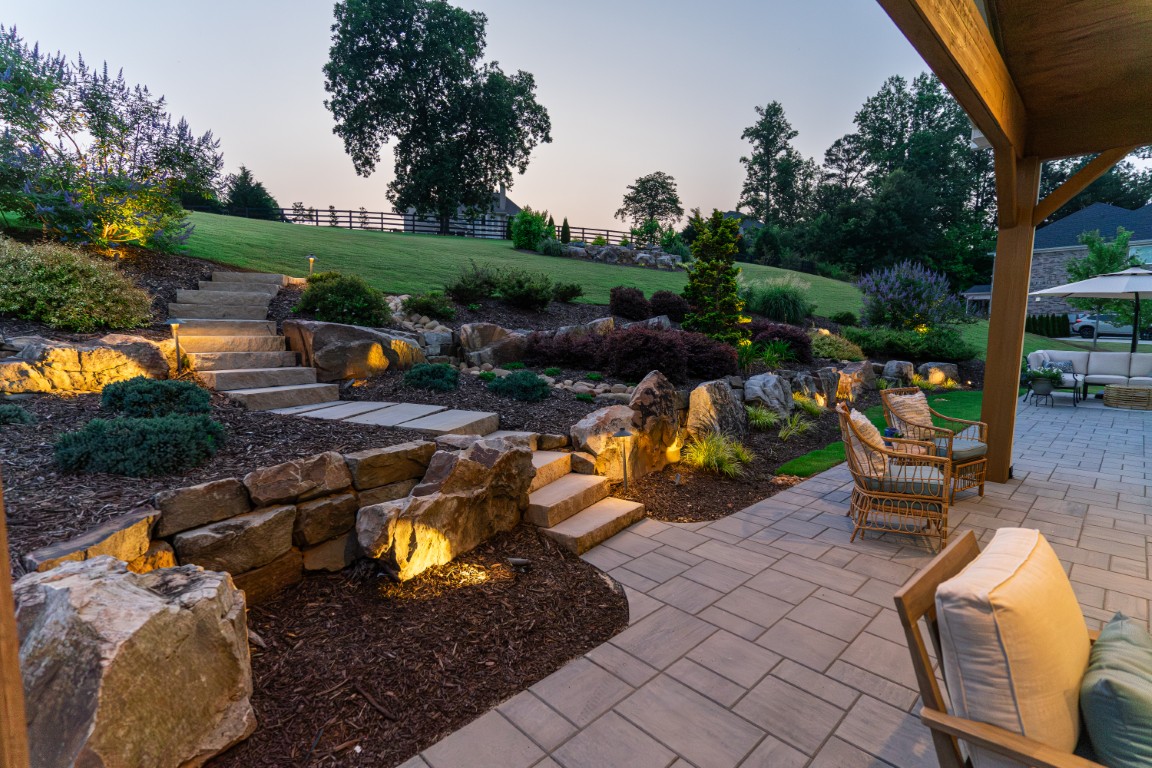 A landscaped backyard at sunset features stone steps, large rocks, outdoor lighting, patio seating, and a paved area near a grassy hill with trees.