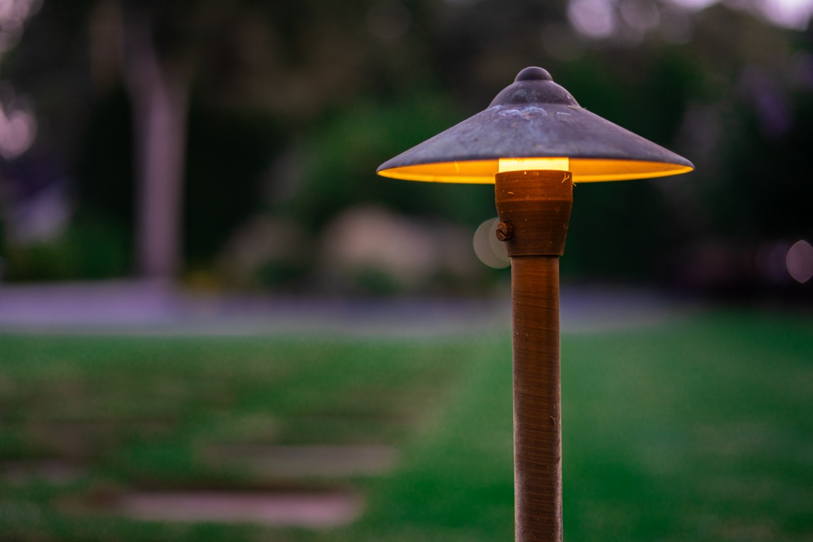 A single garden lamp glows warmly at dusk, with a blurred green lawn and trees in the background.