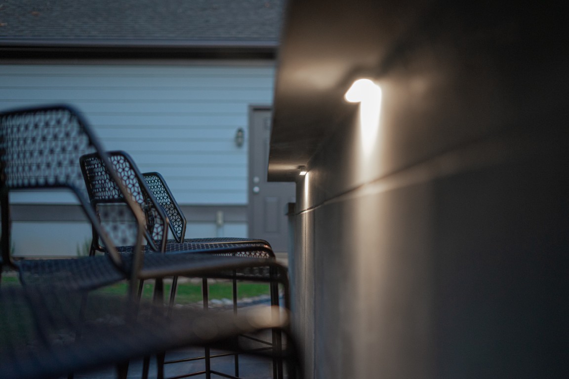 Black metal chairs line an outdoor wall with small lights, in front of a gray house with a door at dusk.