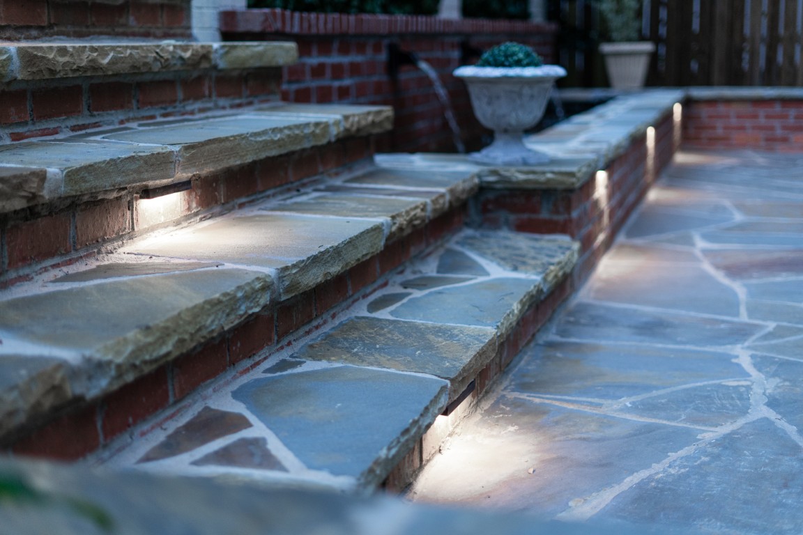 Stone steps with built-in lights lead to a brick wall, with a small potted plant on a step and irregular stone tiles paving the area.